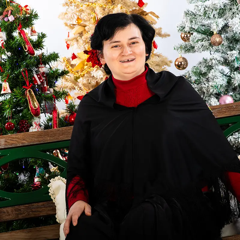 Daniela, un'ospite della comunità residenziale del Centro Papa Giovanni XXIII in posa davanti a 3 alberi di natale riccamente addobbati. Daniela è una donna di 50 anni circa, ha i capelli neri corti ed un volto sorridente e felice. Veste con un maglione nero e rosso.